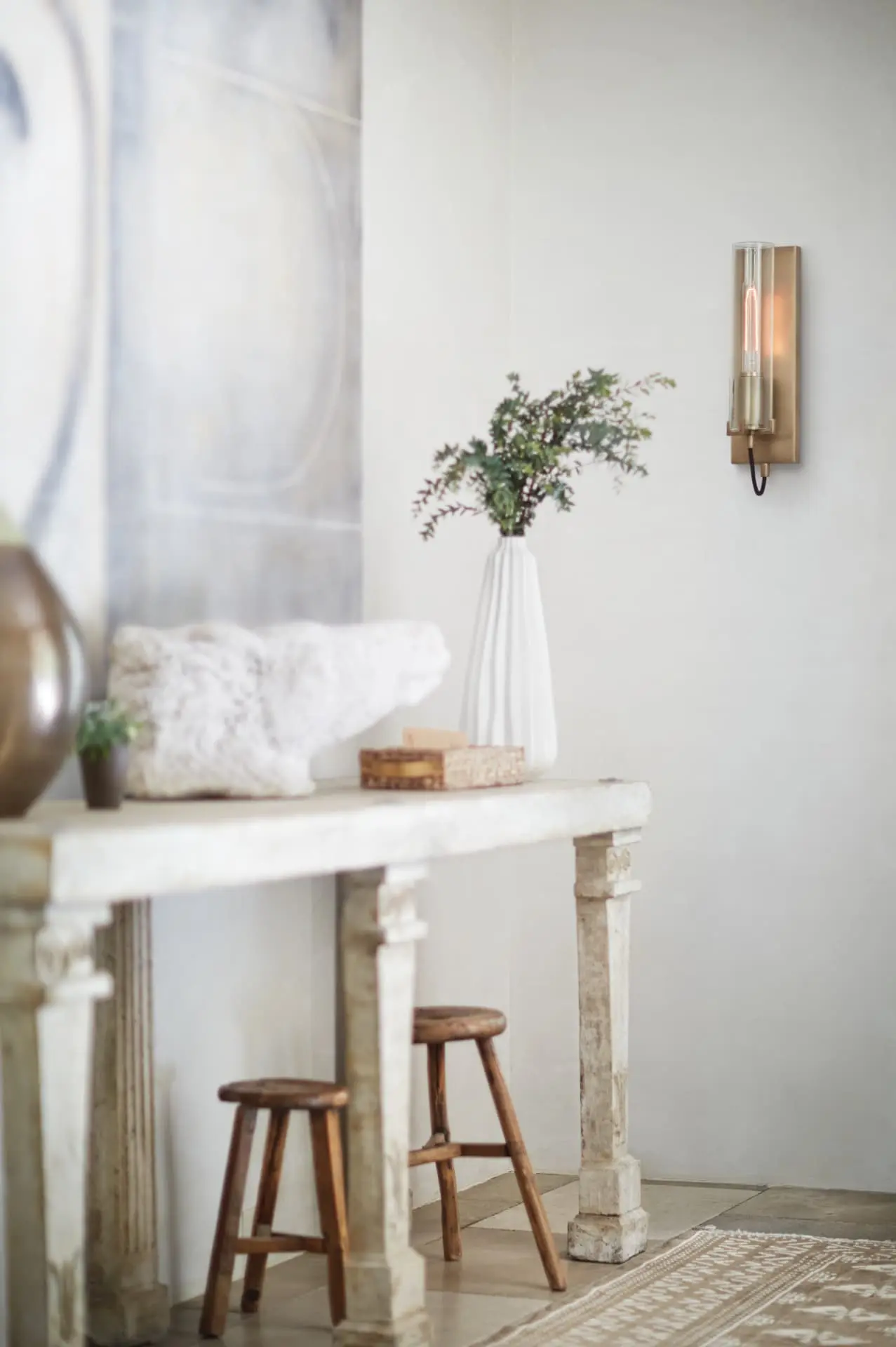 Pale plaster entry with an antique stone console, wood stools and a brass cylinder sconce