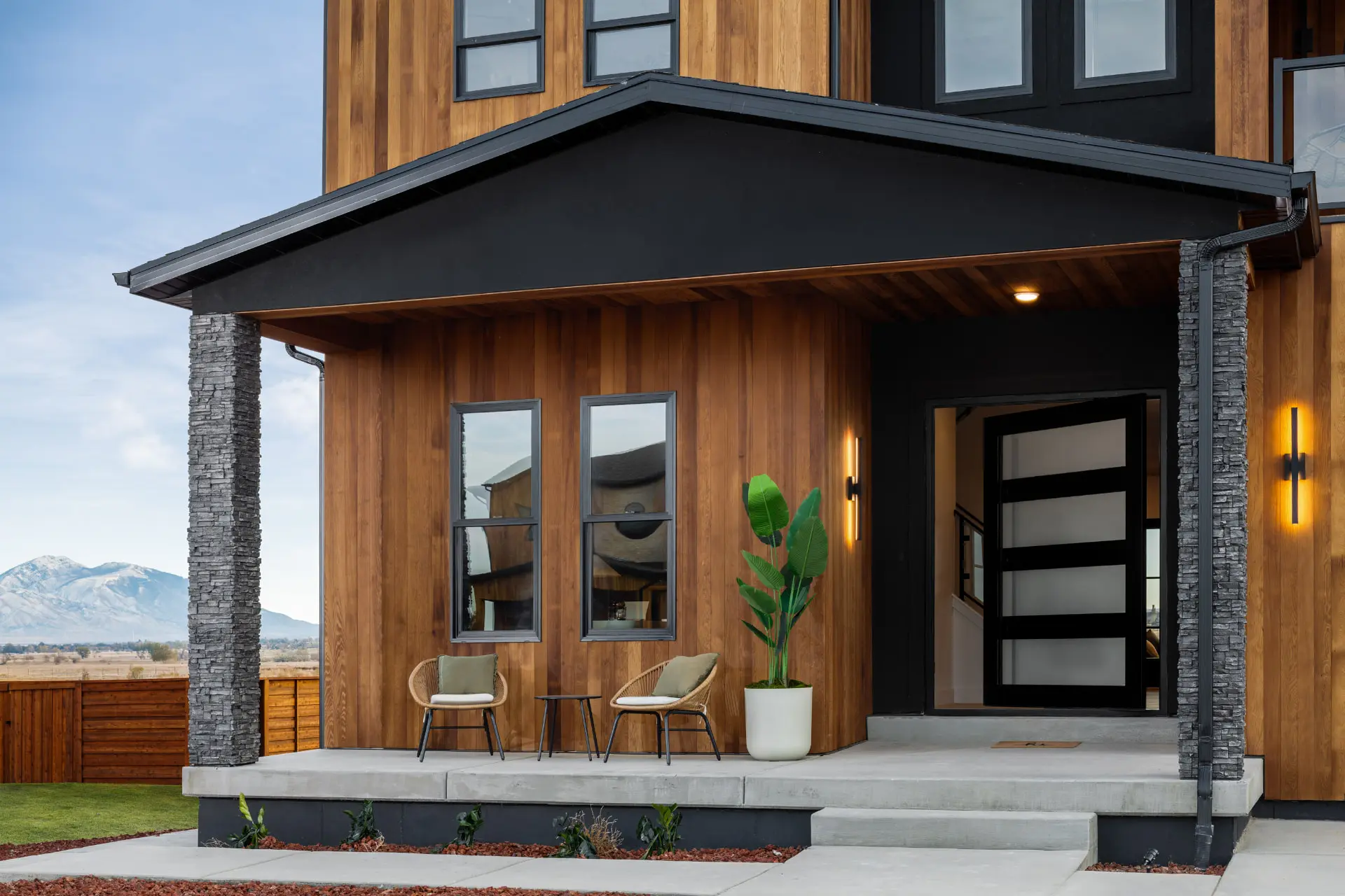 Covered front porch with stacked stone columns and linear LED sconces beside a glass front door