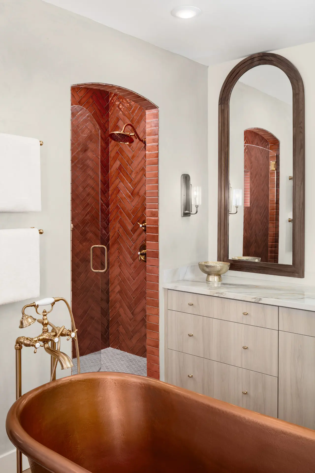 Copper soaking tub beside a terracotta herringbone shower and reeded glass sconce